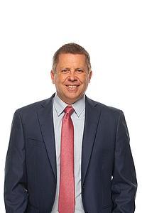 Darren Wilkins stands in front of a white background. He is facing forwards and smiling at the camera.